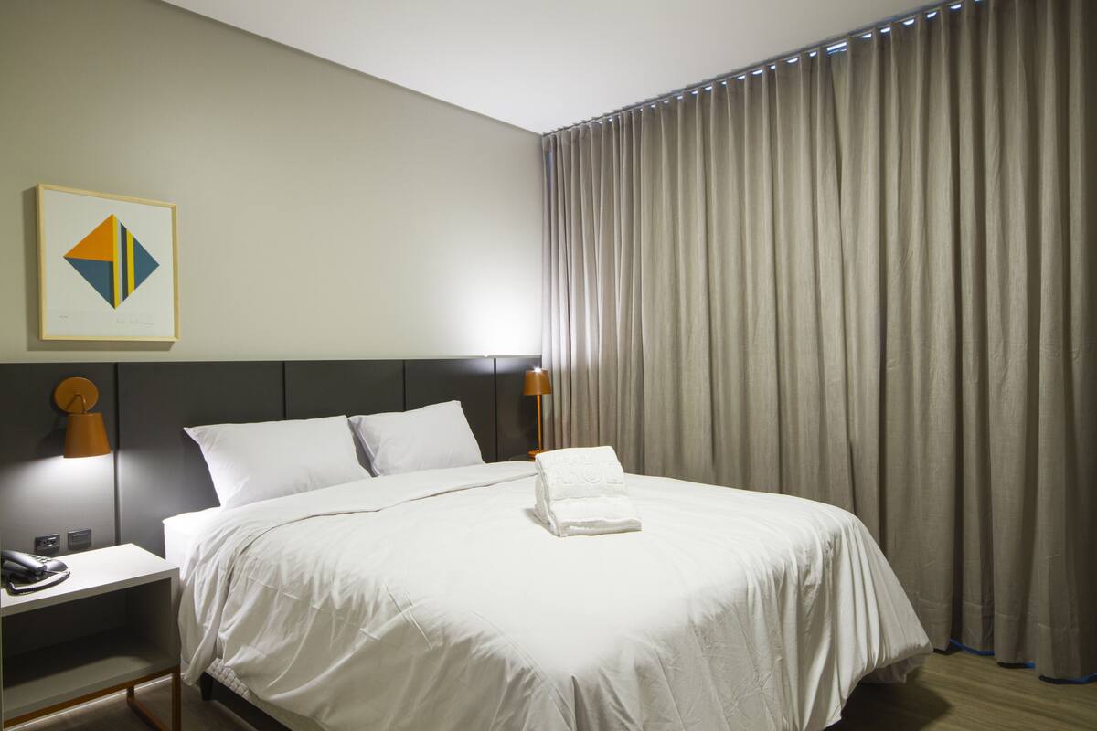 Una habitación de hotel con una cama bien hecha, una lámpara de noche, un cuadro en la pared y una cortina.