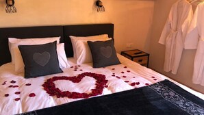 Ein Bett mit Rosenblättern, die in Herzform drapiert sind, zwei Kissen mit Herzmuster und eine schwarze Tagesdecke.