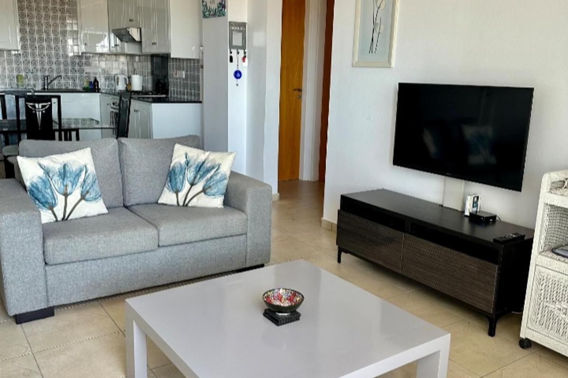 A living room with a grey sofa, a white coffee table, a flat-screen TV, and a wicker chair.