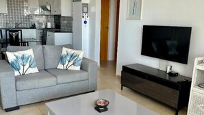 Una sala de estar con un sofá gris, una mesa de centro blanca, un televisor de pantalla plana y una silla de mimbre.
