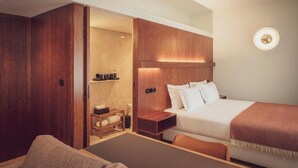 Et moderne hotelværelse med en træskab, en seng med hvide sengetøj, et natbord med en lampe og en lille hylde med toiletartikler.