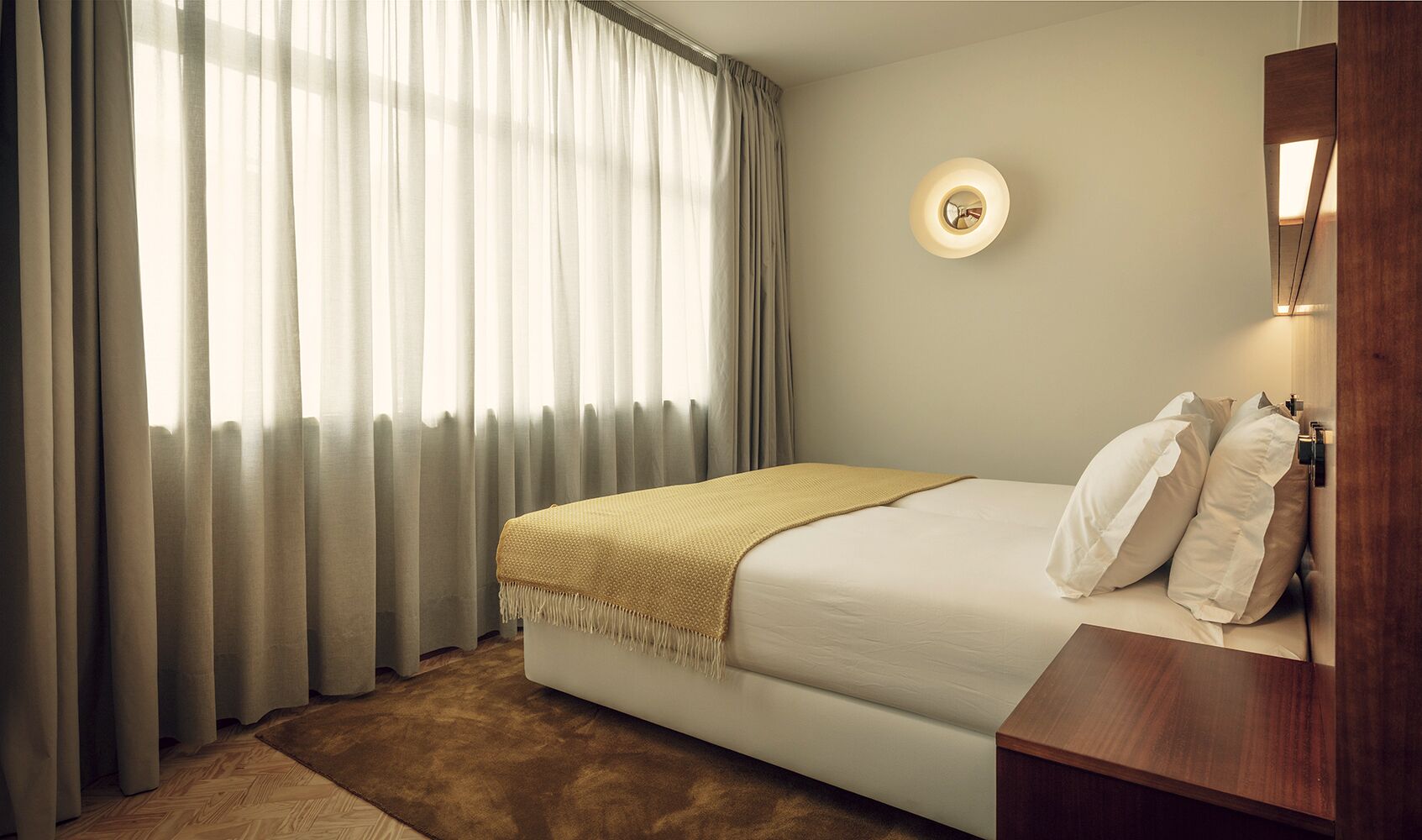 A hotel room with a bed, a bedside table, a wall-mounted light, and a window with curtains.