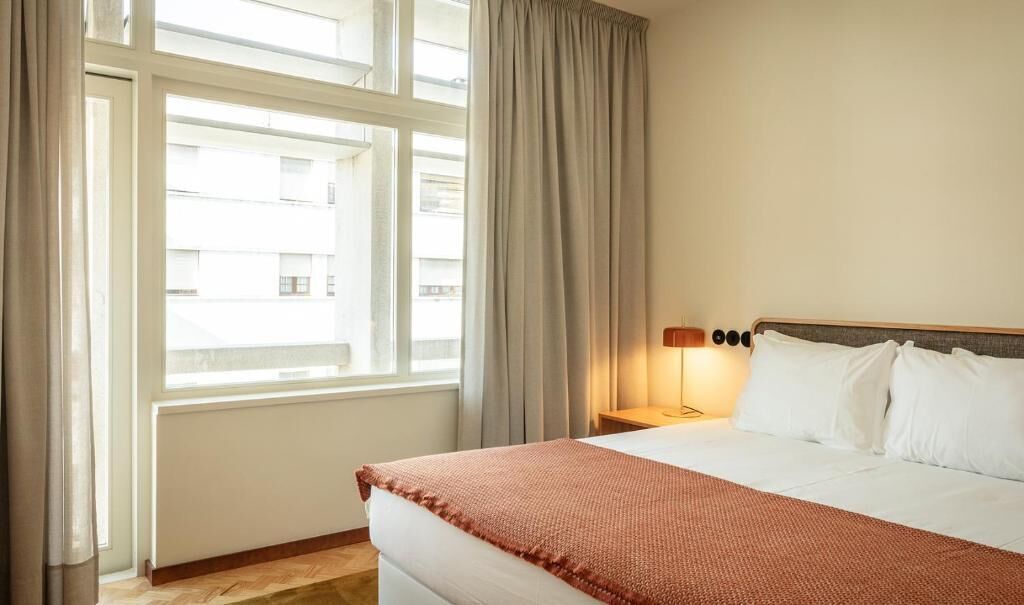 A hotel room with a large window, a bed with white linens, a brown throw blanket, and a nightstand with a lamp.