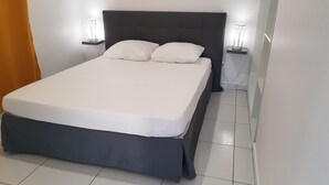 A neatly made bed with white linens and a dark headboard, flanked by two bedside tables with lamps. - The Island Paradise (Baillif)