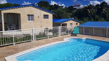 A swimming pool with a white fence, a small building with a blue roof, and a view of a hill with houses.