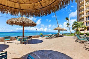 A resort patio with thatched umbrellas, lounge chairs, and a view of the ocean. - Bright Oceanview Studio Steps to Beach West Maui (Lahaina)
