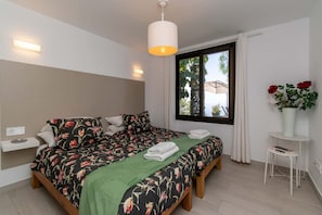 Une chambre à coucher avec un lit, une table de chevet, une fenêtre et une petite table blanche sur laquelle est posé un vase de fleurs.