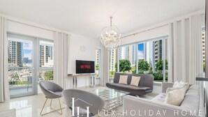 Ein geräumiges Wohnzimmer mit grauem Sofa, einem Glastisch und einem modernen Kronleuchter. Ein Flachbildfernseher ist an der Wand befestigt, und das Zimmer bietet große Fenster mit Blick auf die Stadt.