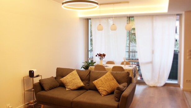 Een moderne woonkamer met een bruine bank, een staande lamp en een eethoek op de achtergrond.