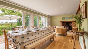 A dining area with a long table set for six, surrounded by wooden chairs, a bench, and a wicker sofa. A fireplace is visible in the background, and the room has large windows offering a view of greenery outside. - Foxhole Cottage,  Framsden, Framlingham (Stowmarket)
