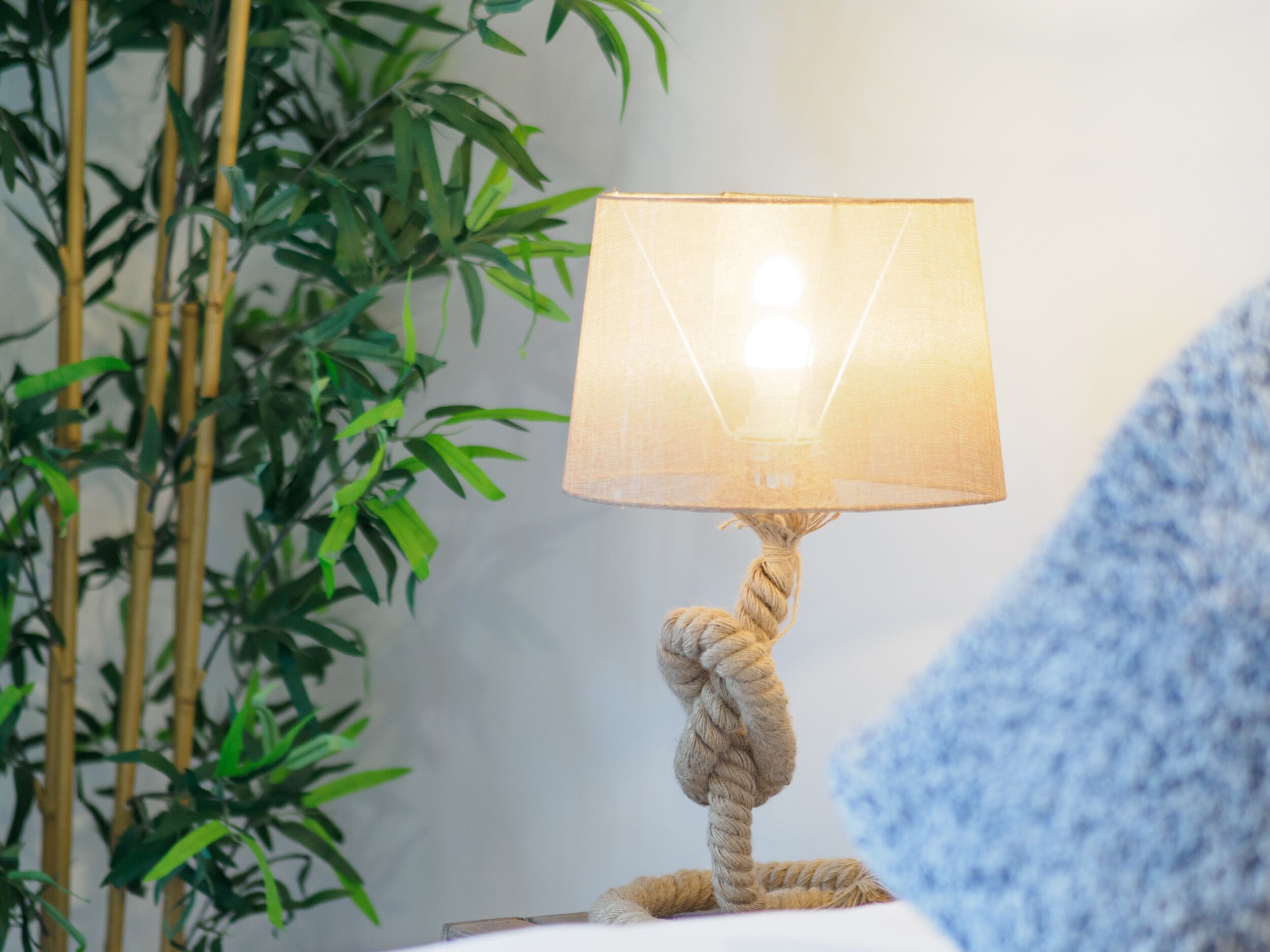 A lamp with a rope base and a fabric shade, placed on a surface with a blue fabric in the foreground.