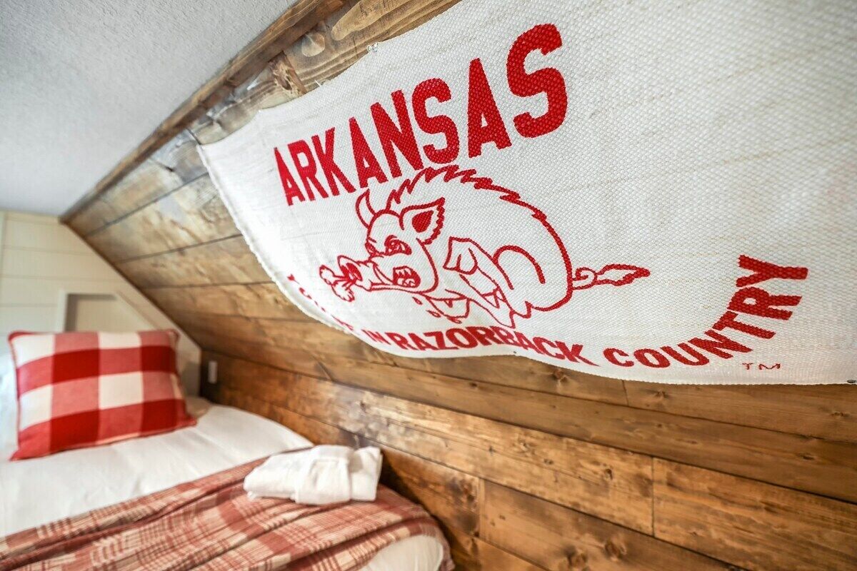 A bed with a checkered pillow and a blanket, a wooden wall, and a banner with "ARKANSAS" and a logo.