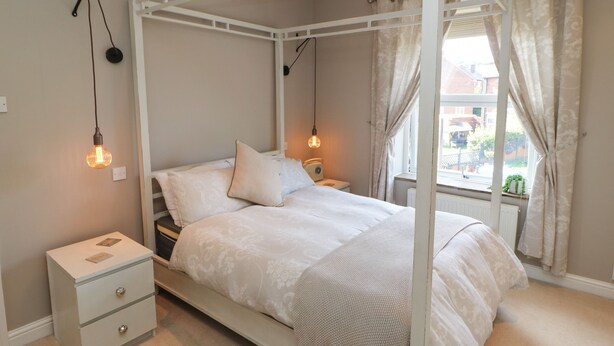 Una cama con dosel y mosquitera blanca, dos mesitas de noche con lámparas, una mesita auxiliar con radio y vistas a la calle a través de una ventana con cortinas.