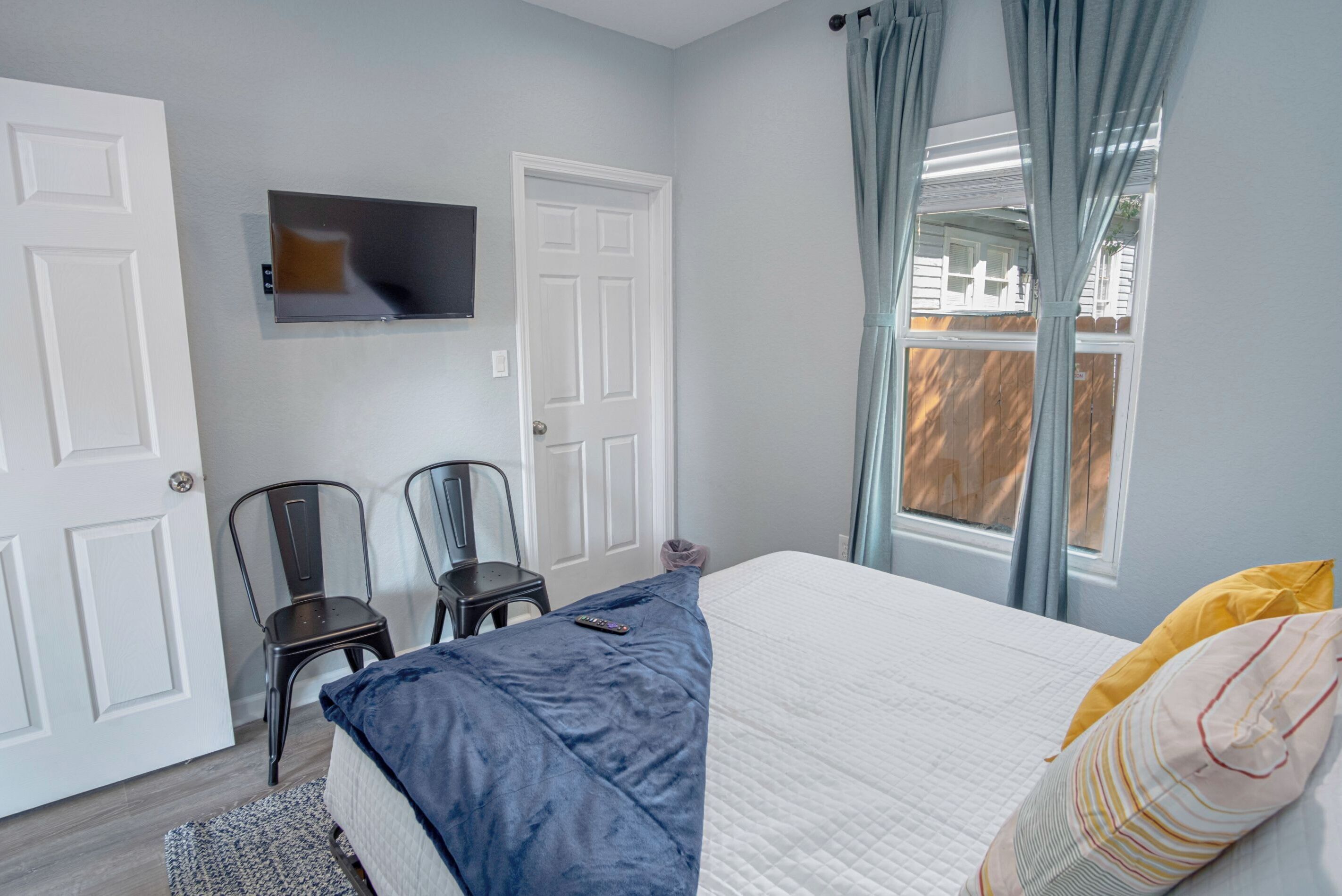 Une chambre à coucher avec un lit, un téléviseur fixé au mur, deux chaises et une fenêtre avec des rideaux.