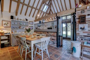 A rustic dining area with a wooden table, white chairs, and a brick wall. - Rooster Barn | East Ruston Cottages (Trunch)