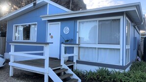 Ein kleines, eingeschossiges Gebäude mit blauer Fassade und weissen Verzierungen. Es hat eine Veranda mit einer Holztreppe und eine Tür mit der Aufschrift 'Blue Shack'.