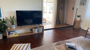 Ein Wohnzimmer mit einem Flachbildfernseher auf einem Holzständer, einem Bücherregal mit Gesellschaftsspielen, einer Topfpflanze und einem Korbtisch.