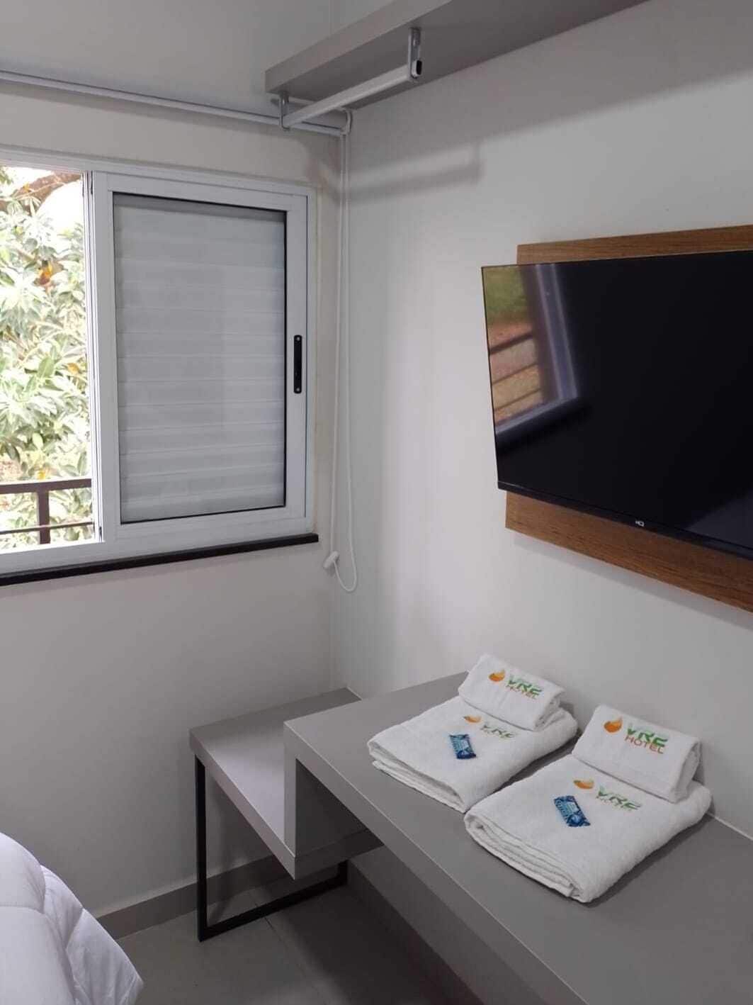 A hotel room with a large window, a flat-screen TV mounted on the wall, a small bedside table, and neatly folded towels with the hotel logo.