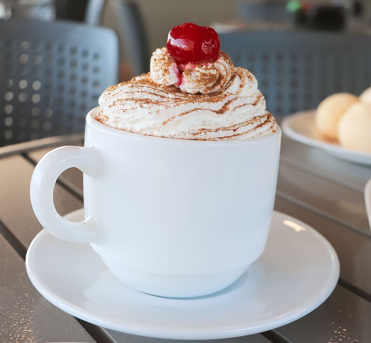 Una taza blanca de café con crema batida y una cereza encima, sobre un platito blanco.