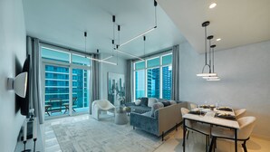 A modern living room with a sofa, dining table, and chairs, large windows with a city view, and a flat-screen TV mounted on the wall. - Maison Privee - Sunrise Bay Tower 2 (Dubai)