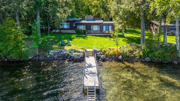 A modern house with a dock extending into a lake, surrounded by dense trees. - 4 Bedroom Waterfront Home With Beatutiful Lake and Mountain Views (Moultonborough)