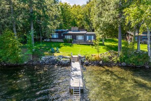 A modern house with a dock extending into a lake, surrounded by dense trees. - 4 Bedroom Waterfront Home With Beatutiful Lake and Mountain Views (Moultonborough)