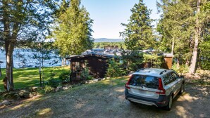 A car parked in a driveway with a wooden cabin and a lake in the background. - 4 Bedroom Waterfront Home With Beatutiful Lake and Mountain Views (Moultonborough)