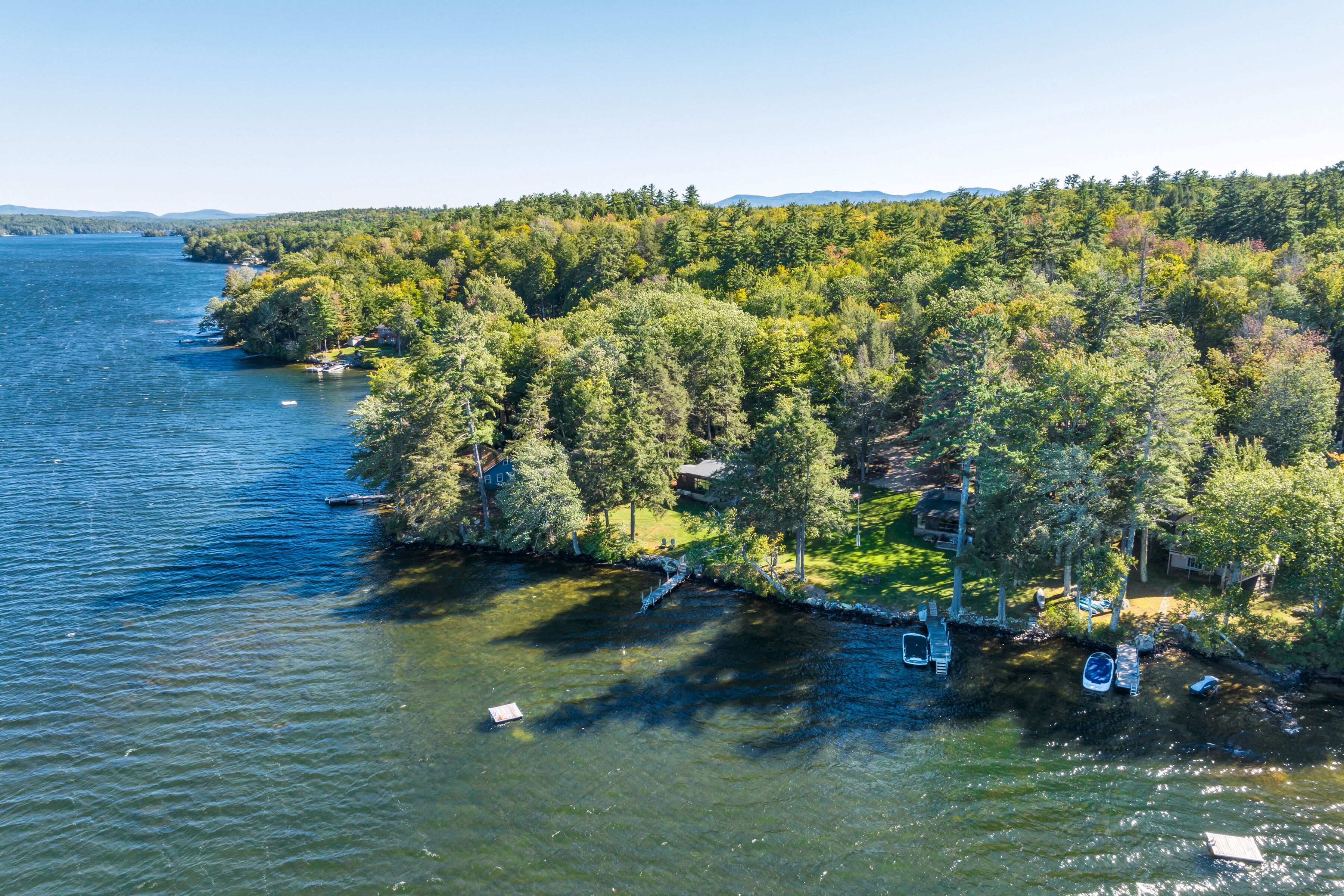 Vista aérea de una propiedad junto al lago con bosque denso, un muelle con barcos y una casa pequeña.