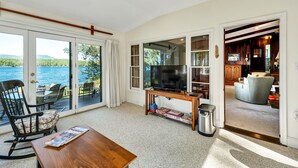 A living room with a TV, a wooden table, a rocking chair, and a view of the water. - 4 Bedroom Waterfront Home With Beatutiful Lake and Mountain Views (Moultonborough)