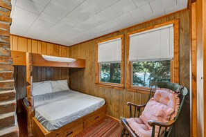 Une chambre dans une cabane en bois, avec un lit superposé, un fauteuil à bascule et deux fenêtres équipées de stores.