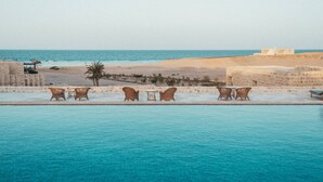 A poolside area with wicker chairs and tables, a sandy beach, and a clear view of the sea. - Wadi Sabarah Lodge (Marsa Alam)