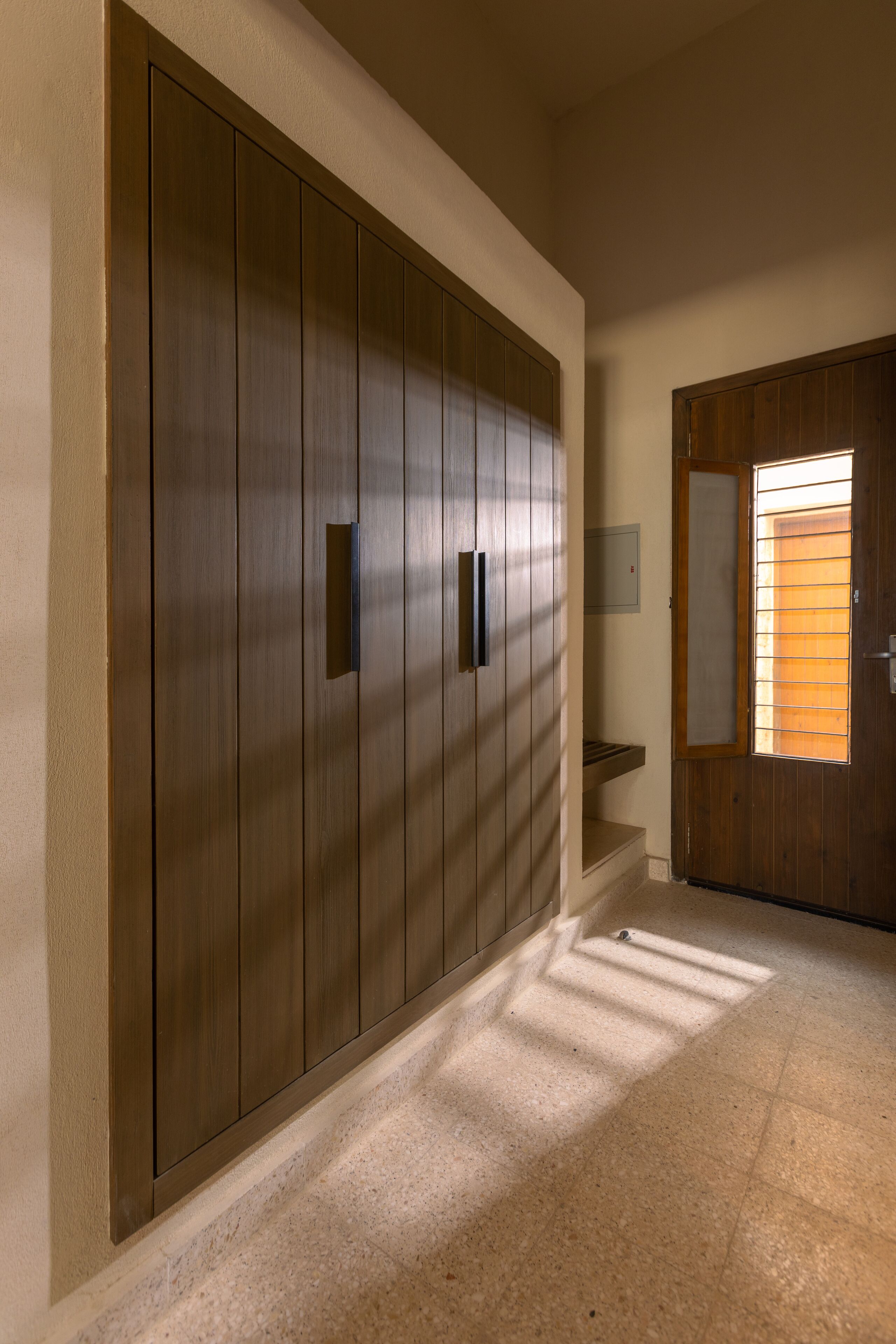 A hallway with a large wooden wardrobe, a small step, and a door with a window.