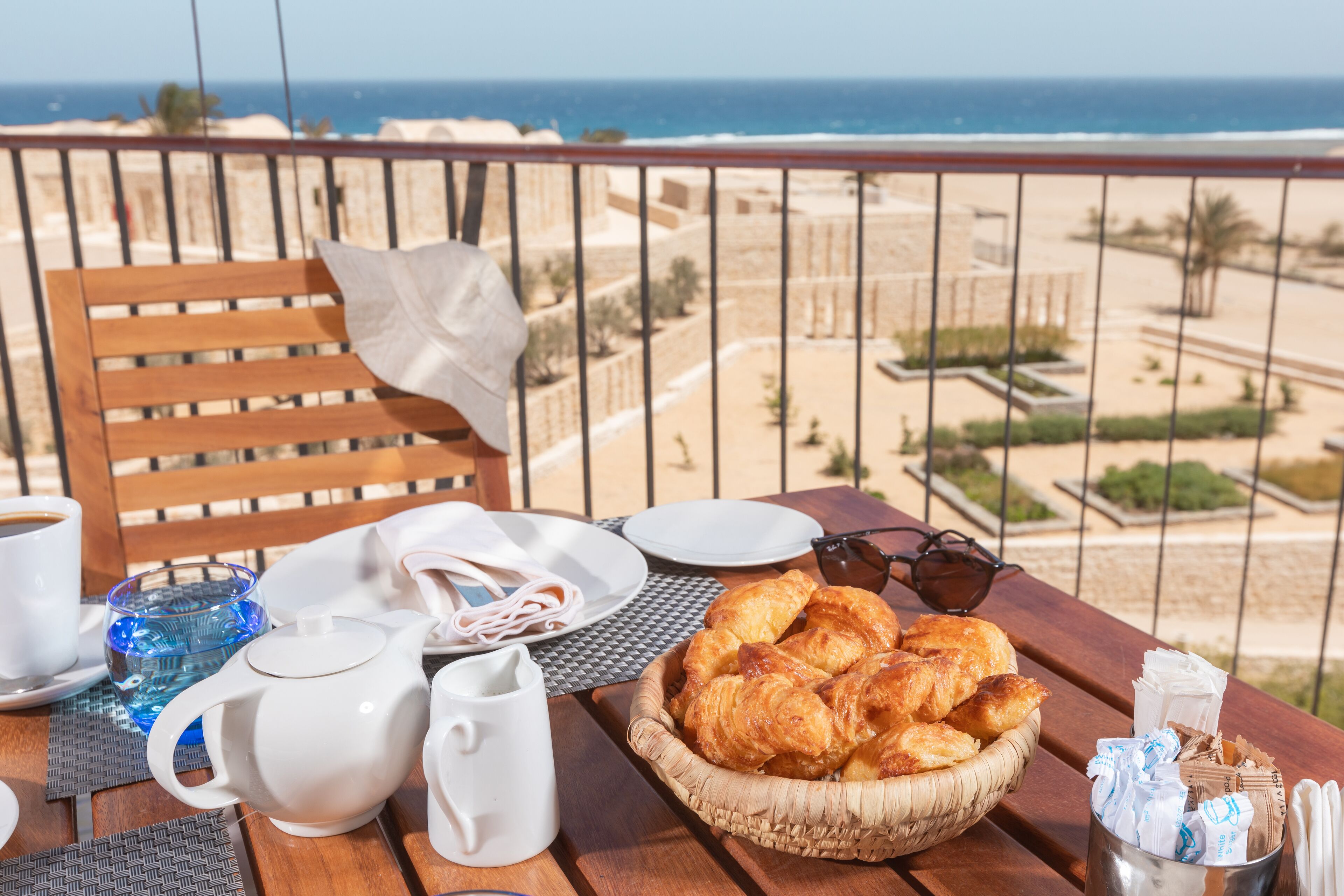 Una mesa de madera con alimentos para el desayuno, como cruasanes, un hervidor de agua y una taza de café, con vista a una playa.
