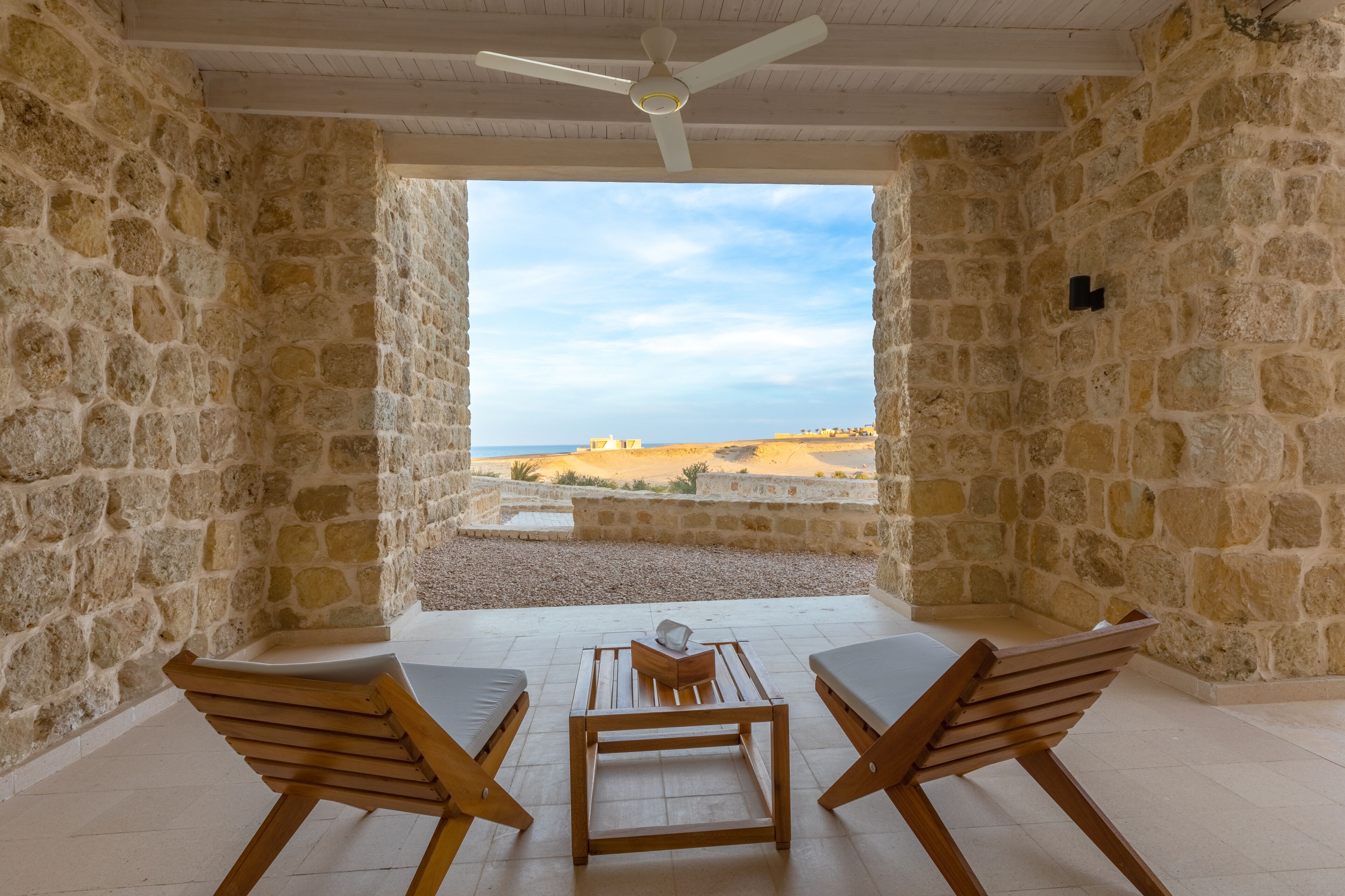 Habitación de paredes de piedra con ventilador de techo, dos sillas de madera, una mesita y vista a la playa.