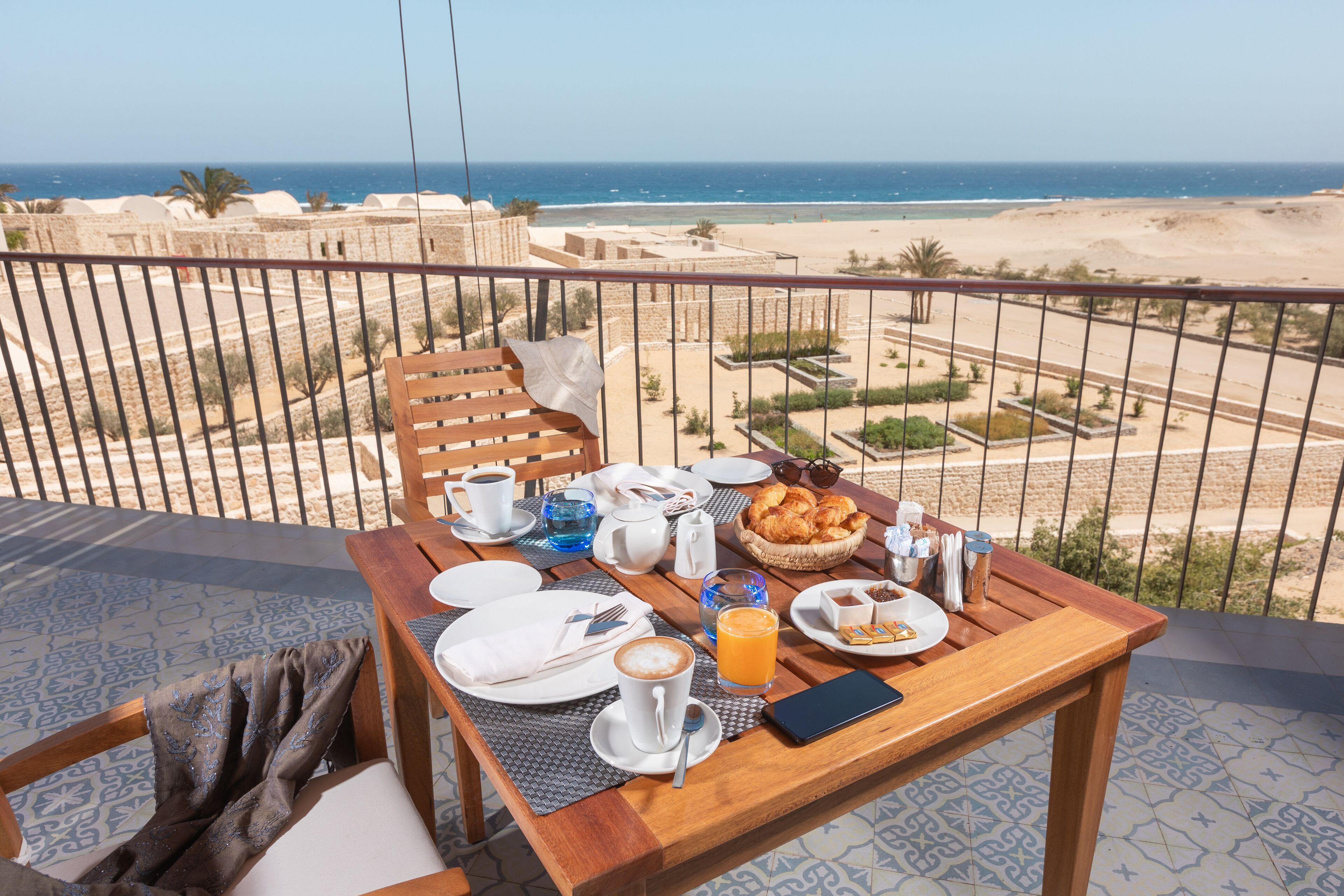 A breakfast table with plates, cups, and a smartphone on a balcony overlooking a beach.