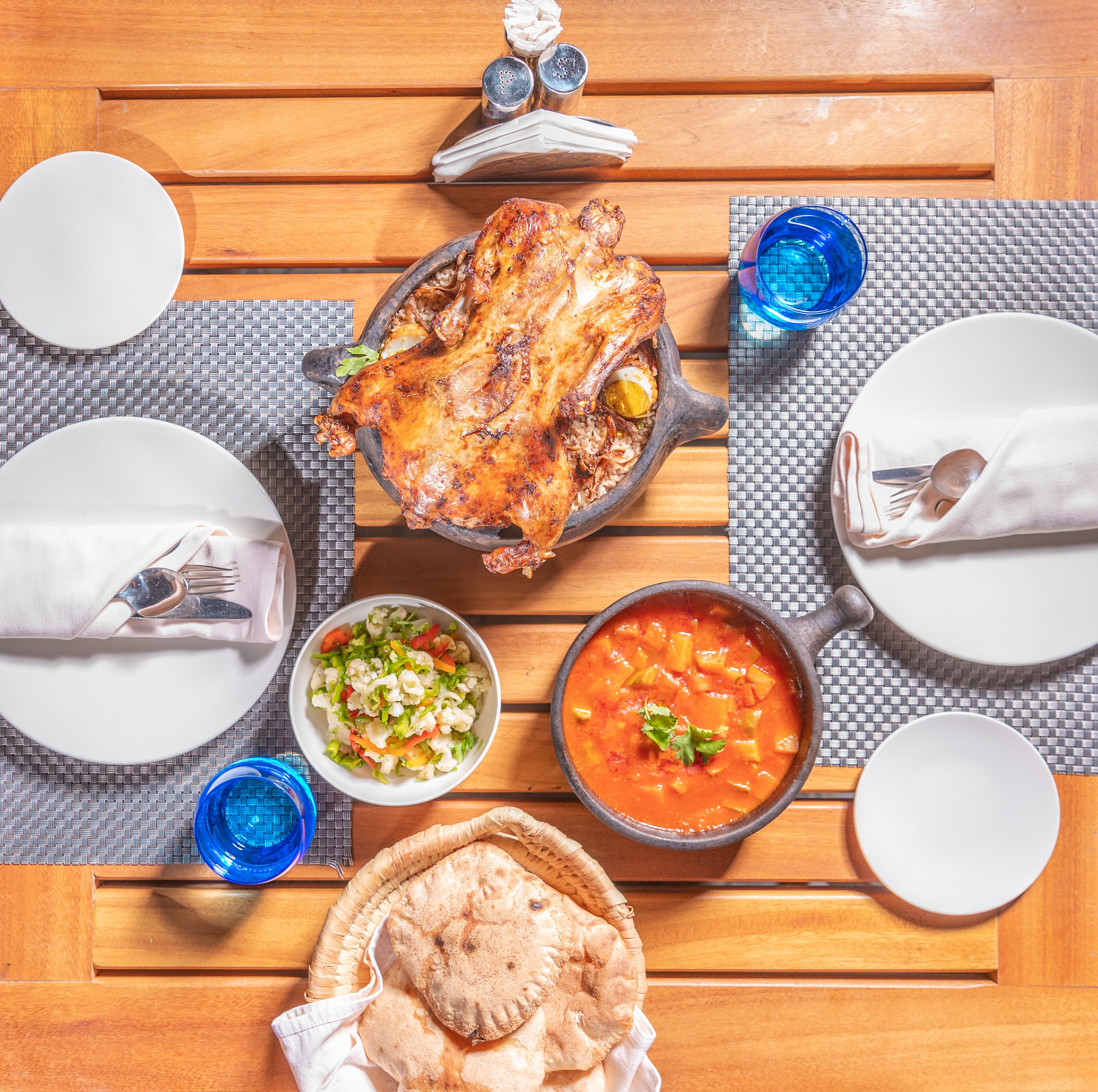 A wooden dining table with a roasted chicken, salad, soup, and flatbread.