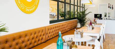 A modern cafe interior with a large yellow logo, wooden tables, white chairs, and a brown cushioned bench.