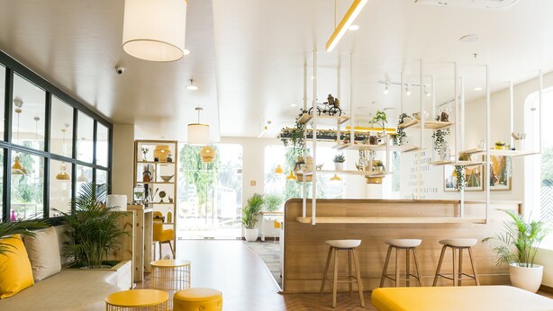 Un intérieur moderne avec un parquet en bois disposé en chevrons, un canapé jaune, un pouf jaune et un coin bar équipé de tabourets et de plantes.