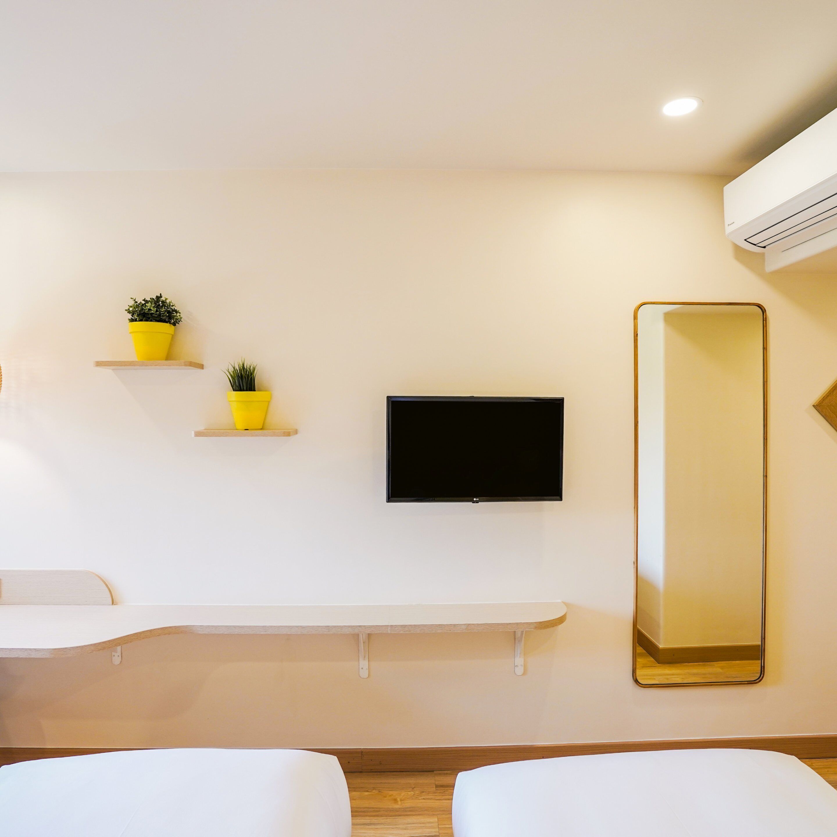 Una moderna camera d'albergo con una TV a schermo piatto fissata al muro, un grande specchio e due piante in vasi gialli su mensole sospese.