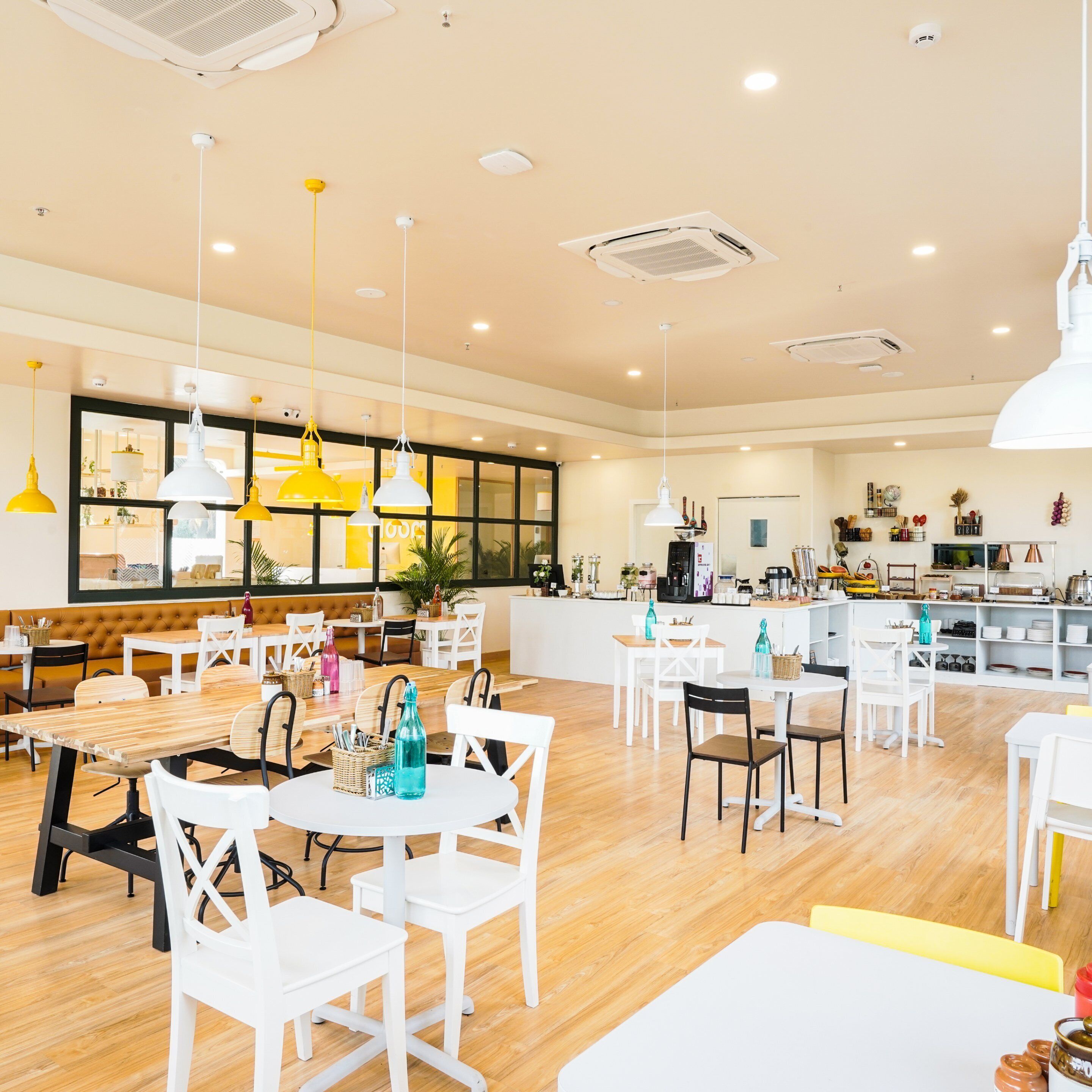 A modern dining area with white tables and chairs, yellow pendant lights, and a kitchen in the background.
