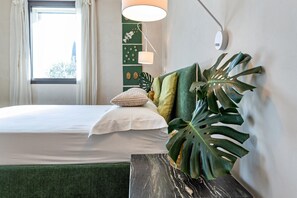 Ein ordentlich bezogenes Bett mit weißen Leinen, ein grün gepolsterter Kopfteil, eine große Zimmerpflanze und eine Wandlampe.