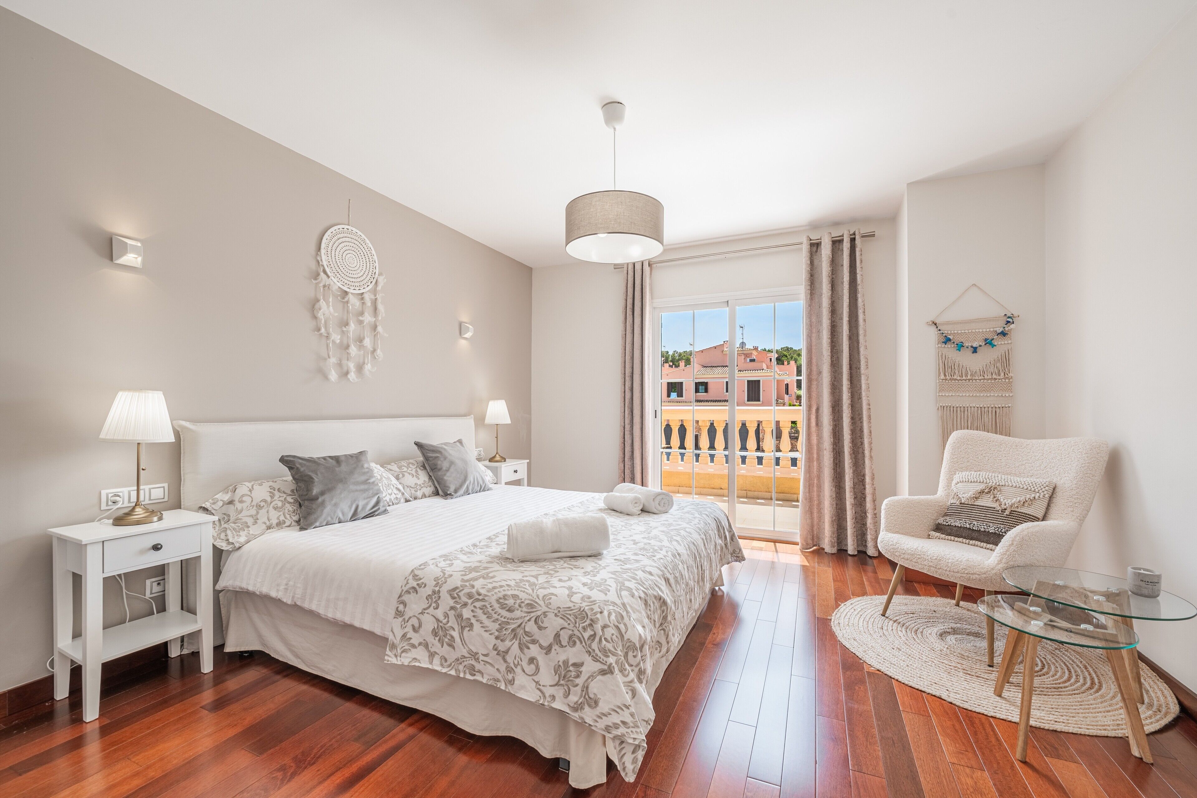 A bedroom with a bed, bedside tables, a chair, a glass table, and a view through a window.