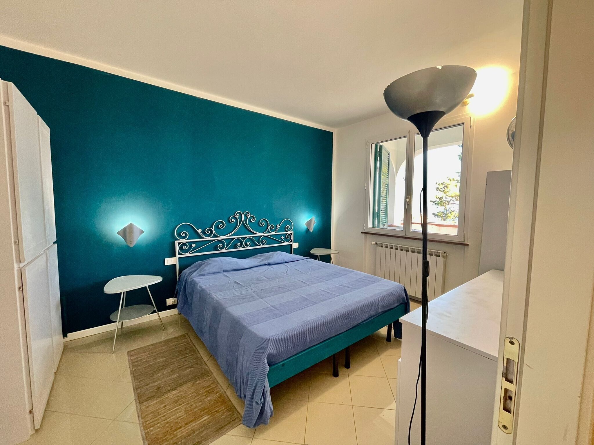Una camera da letto con pareti color acquamarina, un letto con copripiumino blu, un comodino, una lampada da terra e una finestra con vista.