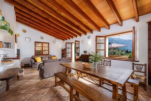 A spacious living area with a wooden ceiling, a large dining table, and a view of mountains through a large window.