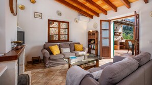 A living room with a sofa, a glass coffee table, a wooden bookshelf, and a view of an outdoor patio. - Holiday Home 'Can Bordoy' with Mountain View, Pool, Garden & Terrace (FORNALUTX)