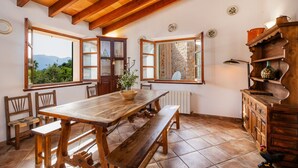 A wooden dining table with benches, a wooden cabinet, and a view of the outdoors through large windows. - Holiday Home 'Can Bordoy' with Mountain View, Pool, Garden & Terrace (FORNALUTX)