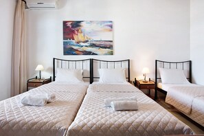 A hotel room with three beds, a painting of a sailboat, and a window with curtains.