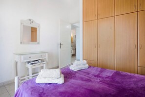 A bedroom with a purple bedspread, a white dresser with a mirror, wooden wardrobe, and a glass-topped table.