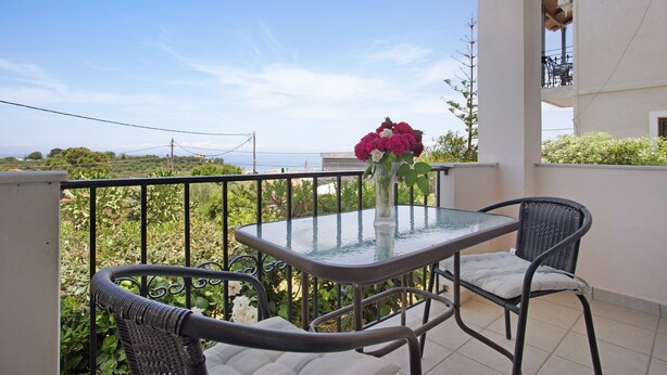 Un balcón con una mesa de vidrio, dos sillas de mimbre, un florero con flores y vista al mar.