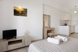 A hotel room with a bed, a TV, a desk with a mirror, a kitchenette, and a painting on the wall.
